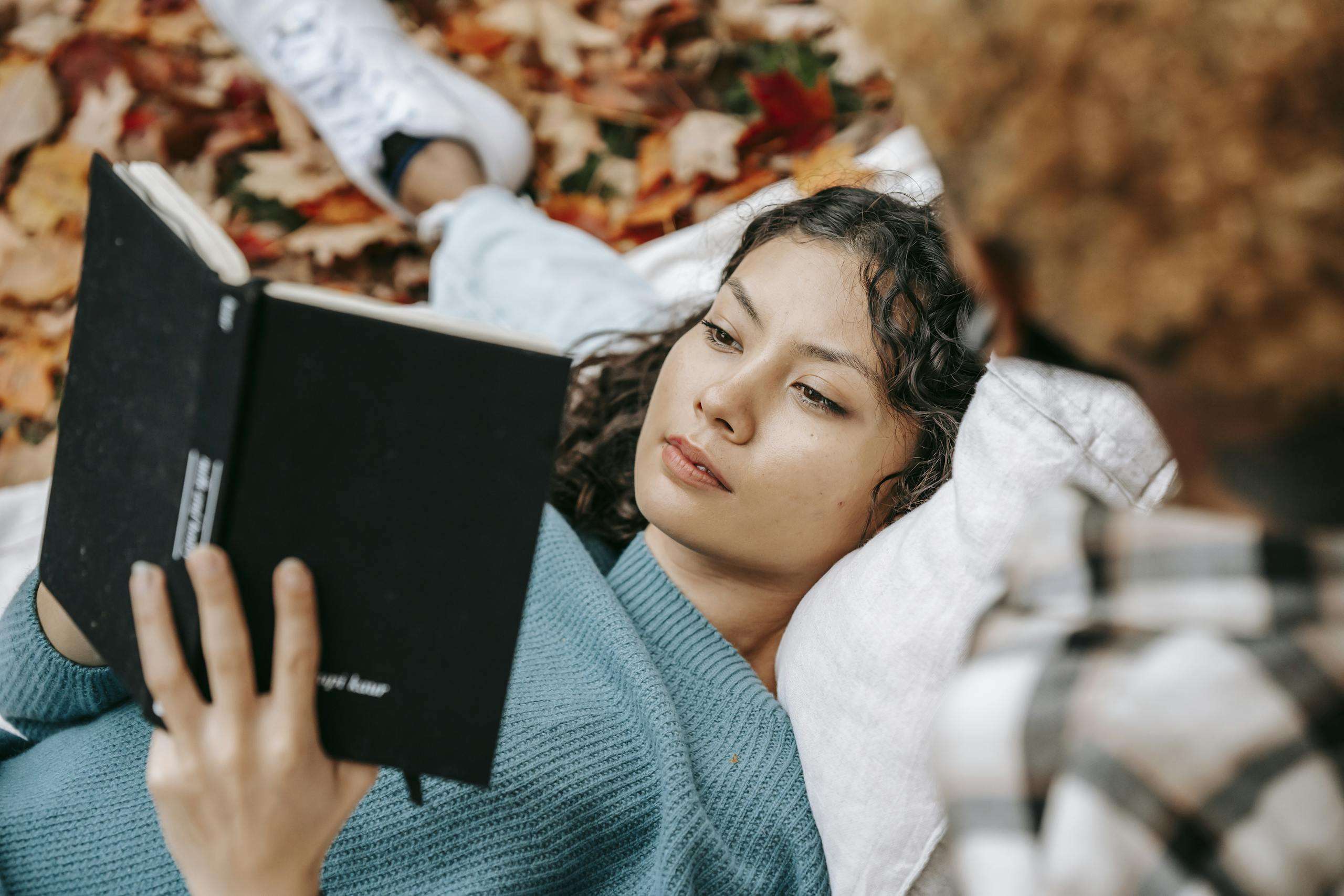 From above of pensive Hispanic female reading book while lying on lap of crop person while spending free time together in autumn nature